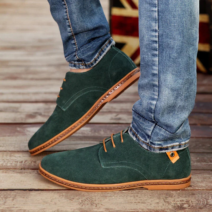 Green suede casual men's shoes with tan soles and laces worn with blue jeans on wooden floor