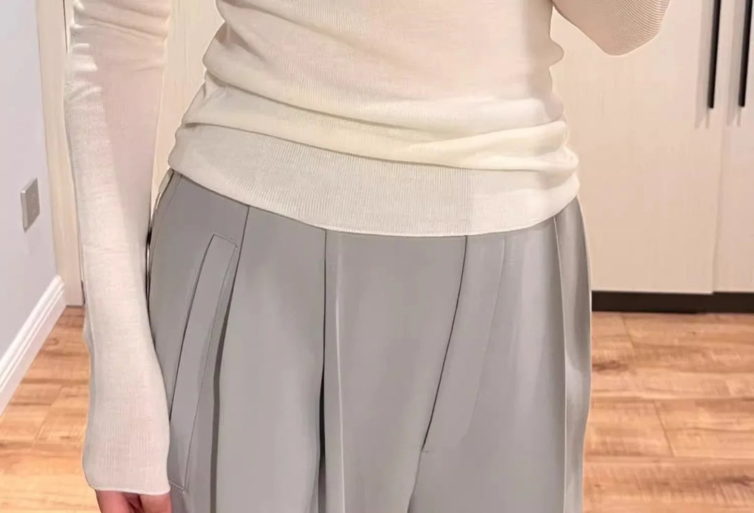 Person wearing white ribbed long sleeve top and light gray pleated pants in wooden indoor setting