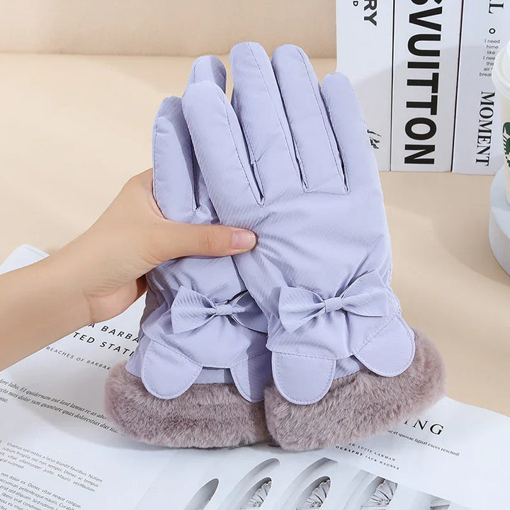 Pair of light purple winter gloves with bows and fluffy cuffs held by hand on a beige surface