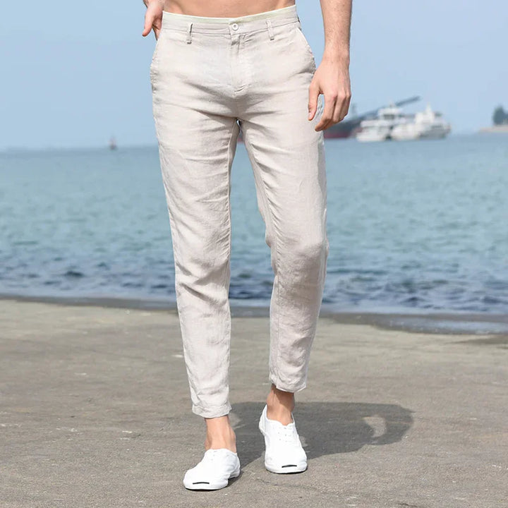 Man wearing beige linen pants and white sneakers walking on seaside pavement with boats in background