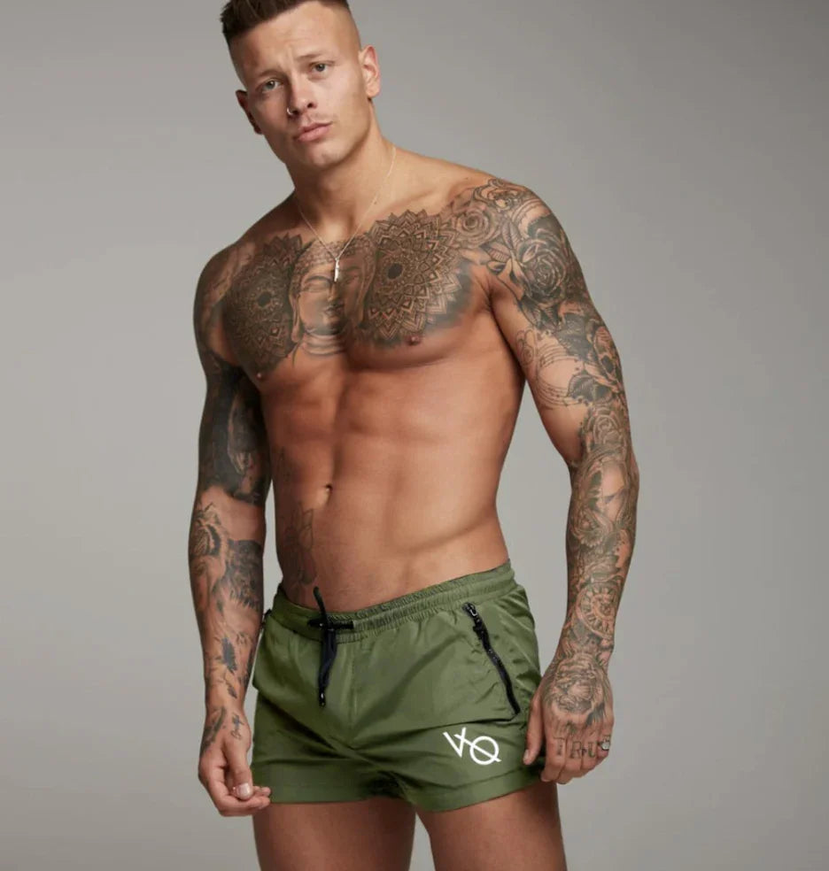 Muscular tattooed man wearing olive green swim shorts with black zipper pockets against gray background