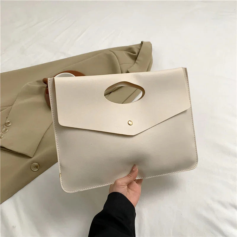 Hand holding a minimalist beige leather clutch bag with a cutout handle, placed over a beige blazer