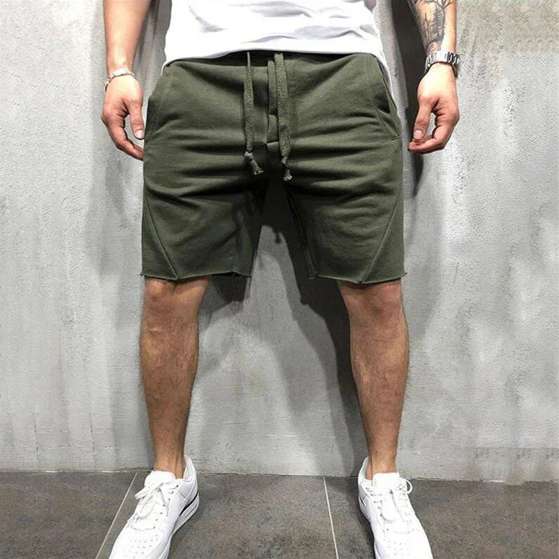 Man wearing olive green casual drawstring shorts and white sneakers against a gray textured wall