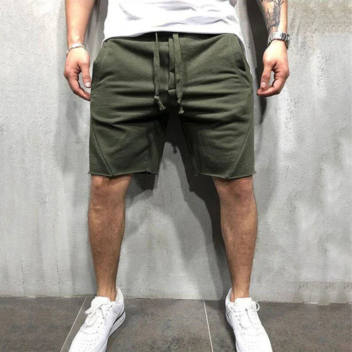 Man wearing olive green casual drawstring shorts and white sneakers against a gray textured wall