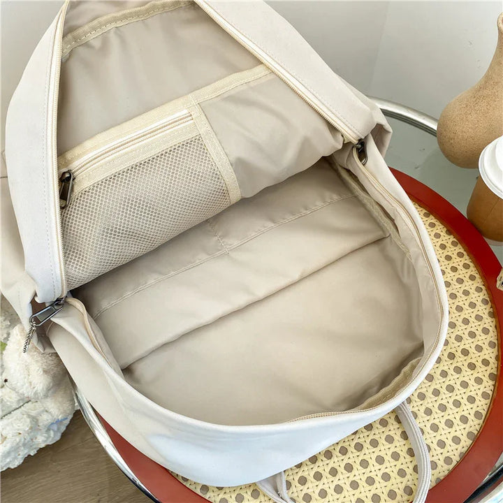 Beige backpack open to show interior with mesh zippered pocket, placed on woven chair with table and coffee cup nearby