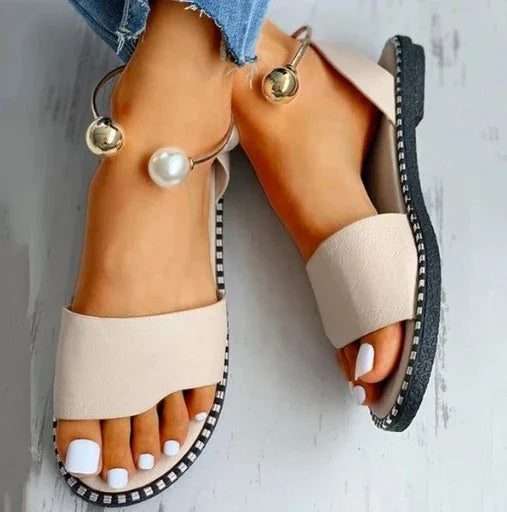 Beige open-toe flat sandals with black sole and white nail polish on feet, paired with ripped blue jeans
