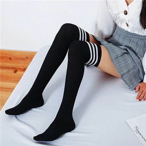 woman wearing black thigh-high socks with white stripes, plaid skirt, and white top sitting on bed