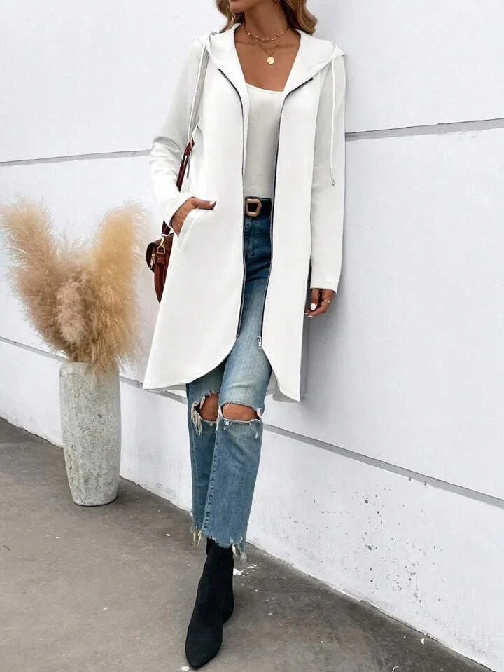 Woman wearing white hooded long coat with zip, ripped jeans, black boots, and layered necklaces near pampas grass vase