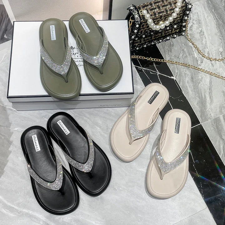 Three pairs of glittery rhinestone flip-flops in olive green, black, and beige on marble floor with a tweed handbag