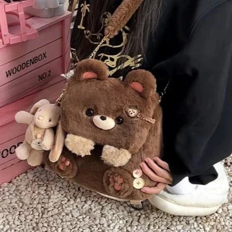 Plush brown bear handbag with buttons and keychain, held by person in black sleeve seated on gravel