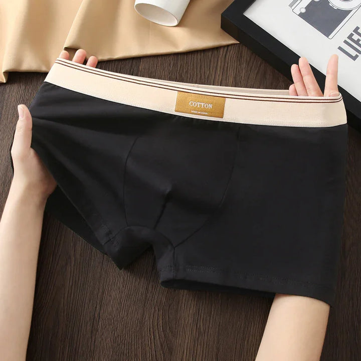 Black cotton men's boxer briefs with beige waistband and label, held stretched on wooden surface