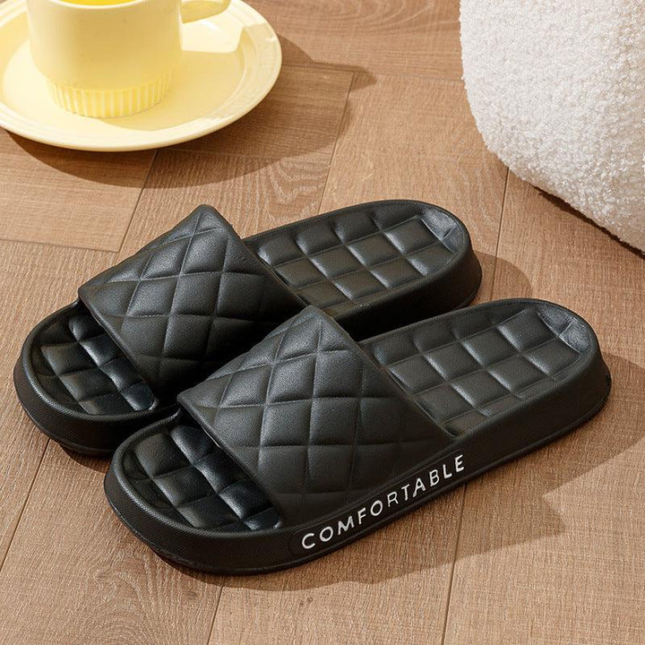 Black quilted cushioned slide sandals with the word Comfortable on the side, placed on wooden floor