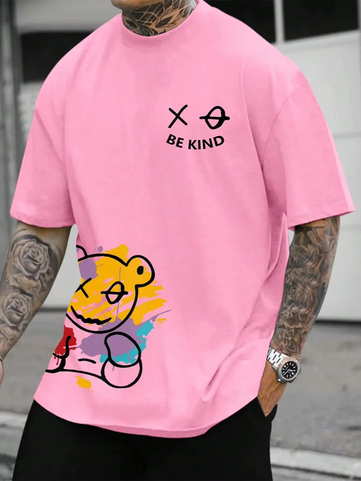 Man wearing pink t-shirt with colorful teddy bear graphic and 'Be Kind' text, tattooed arms