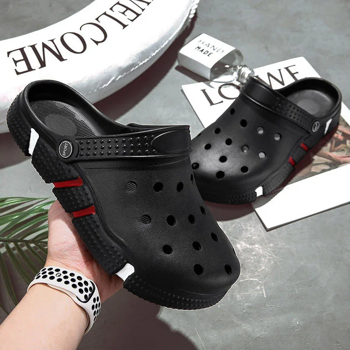 Black slip-on clogs with ventilation holes and red and white sole accents on gray surface