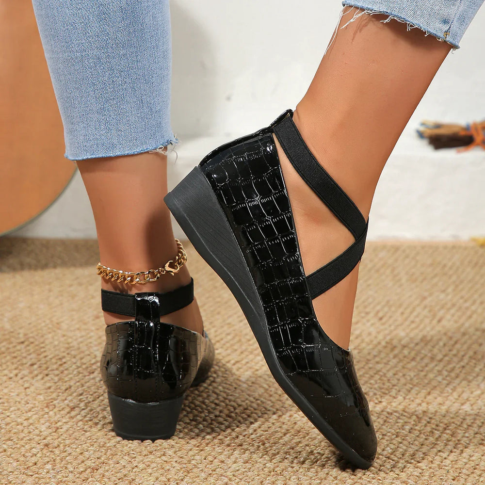 Black patent leather wedge shoes with crisscross elastic straps on feet wearing blue jeans