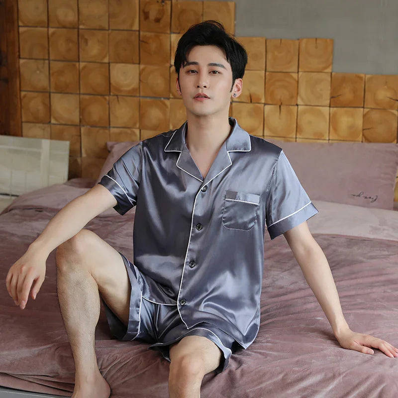 Man sitting on bed wearing shiny grey satin short-sleeve pajama set with white piping