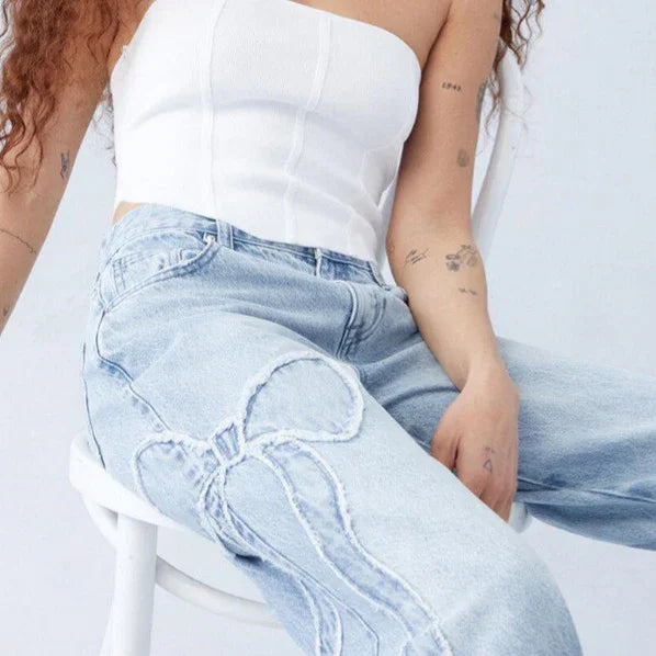 Woman wearing light blue jeans with stitched butterfly design and white tube top sitting on white chair