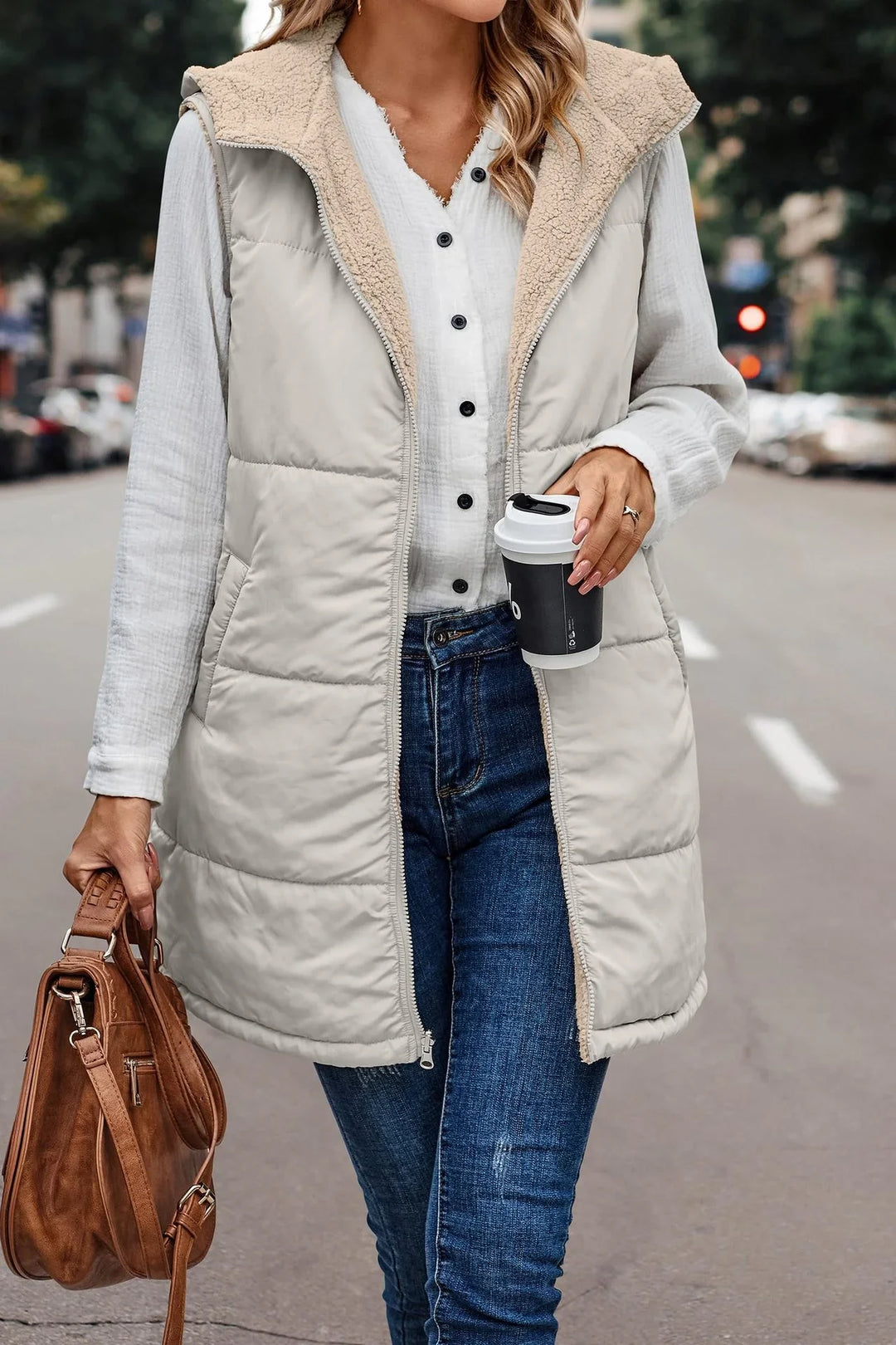 Woman wearing beige quilted hooded vest with sherpa lining, white button shirt, blue jeans, holding coffee cup and brown leather bag