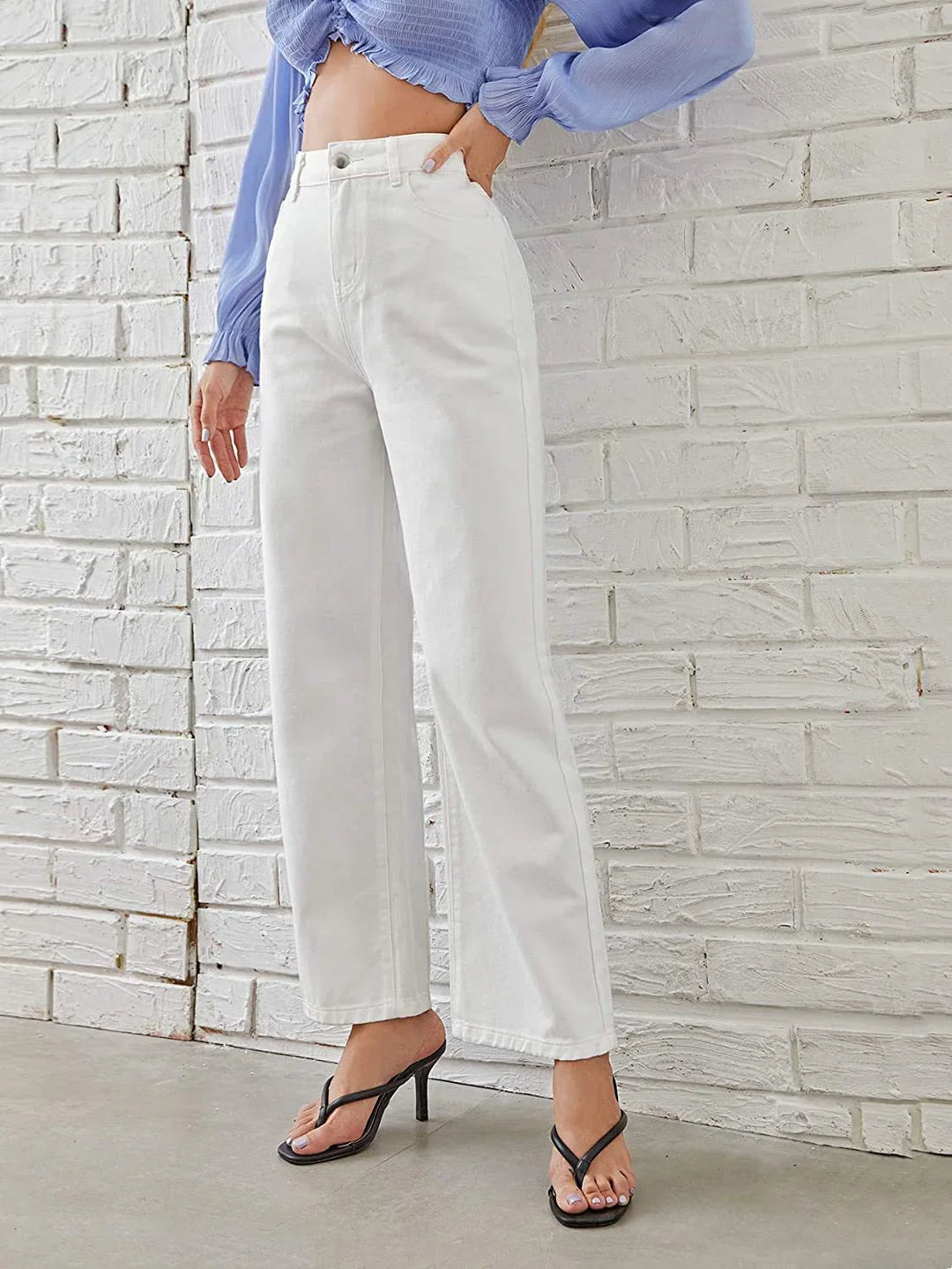 Woman wearing high-waisted white straight-leg jeans and black heeled sandals against white brick wall