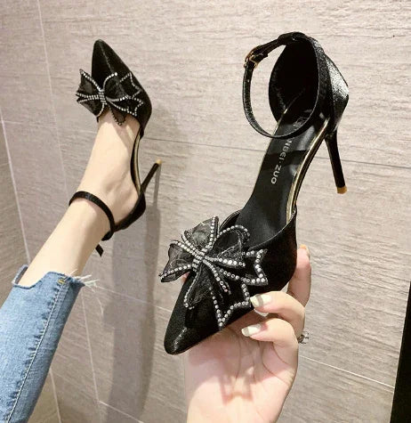 Black high heel shoes with ankle straps and large rhinestone bow decoration, worn with blue jeans