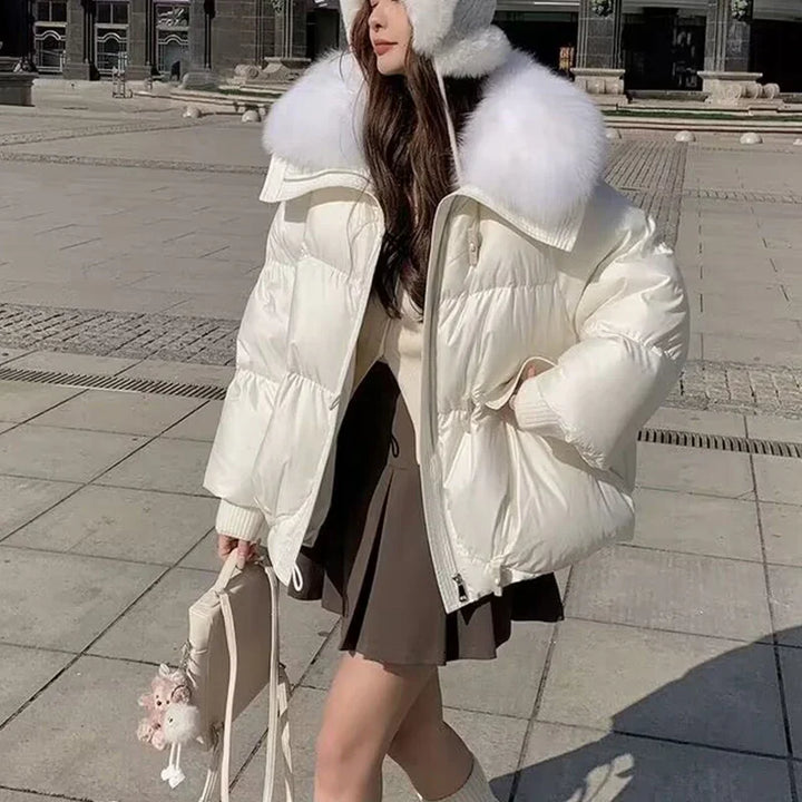 Woman wearing white puffer jacket with fur collar and earmuffs, brown skirt, holding beige bag outdoors