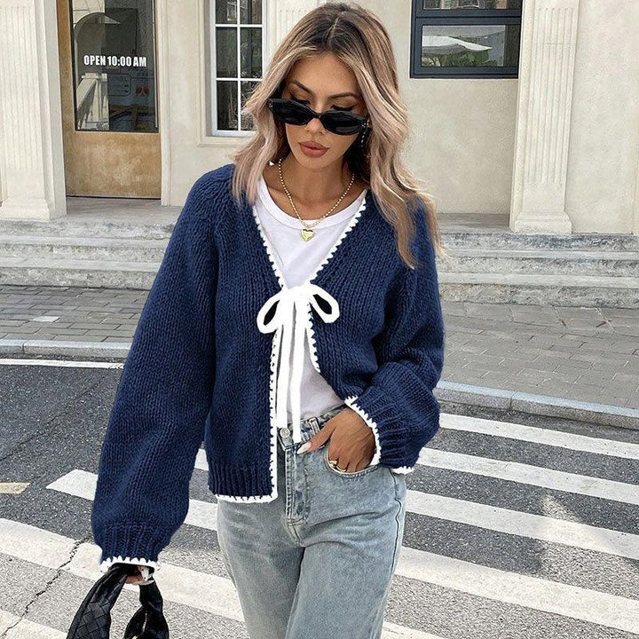 Woman wearing navy knit cardigan with white trim over white top, light denim jeans, black sunglasses, and gold heart necklace on city street