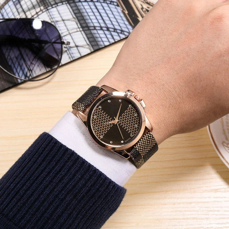Close-up of wrist wearing rose gold watch with black patterned dial and strap, next to sunglasses on wooden table