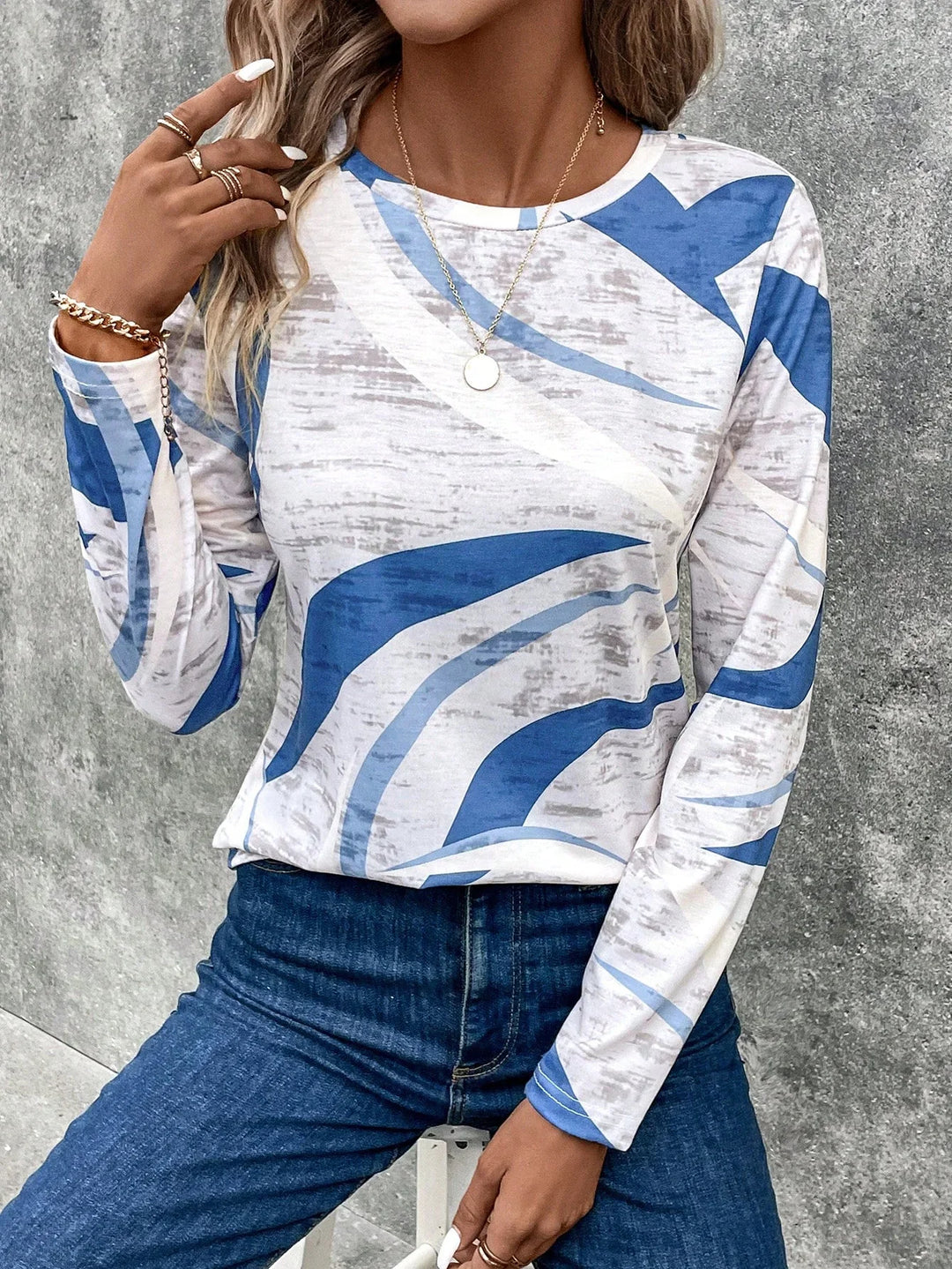 Woman wearing abstract blue and white long sleeve top with gold jewelry and blue jeans against gray wall