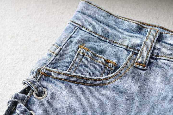 Close-up of light blue denim jeans pocket with stitching and side lace-up detail on white surface