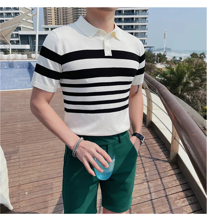 Man wearing white polo shirt with black horizontal stripes and green shorts holding a blue glass on wooden deck