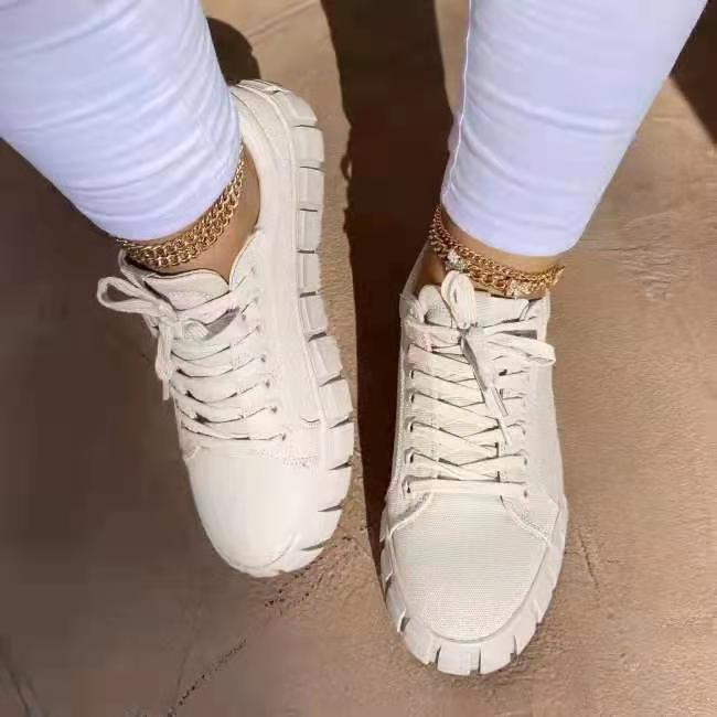 White chunky sneakers worn with white pants and gold chain anklets on a brown floor