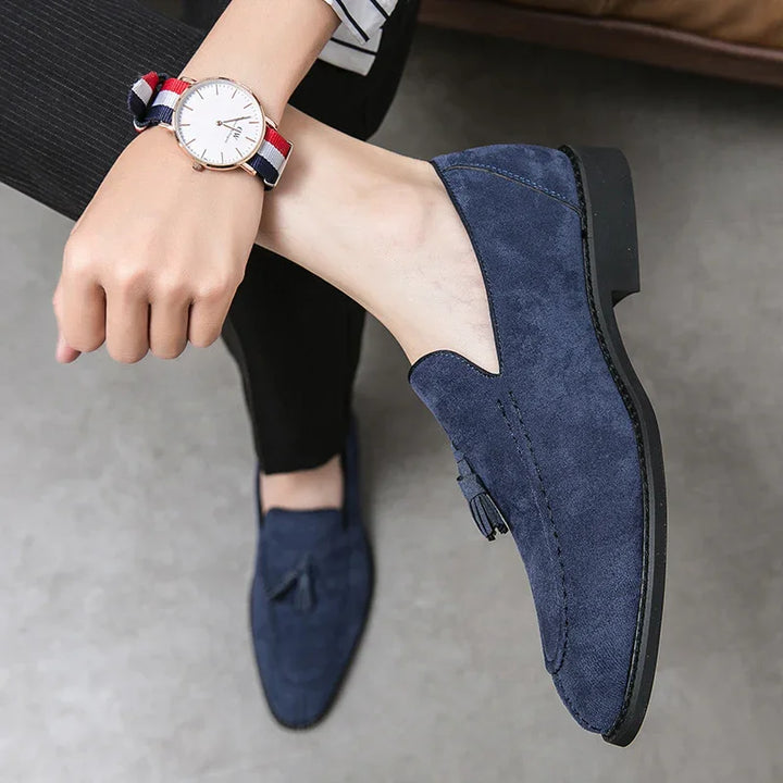 Close-up of navy blue suede tassel loafers worn with black pants on a concrete floor