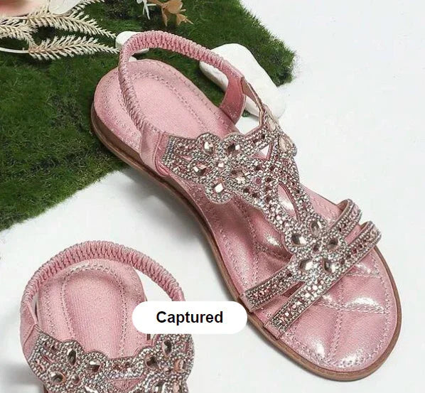 Pink women's sandals with jeweled floral straps on white surface and green moss background