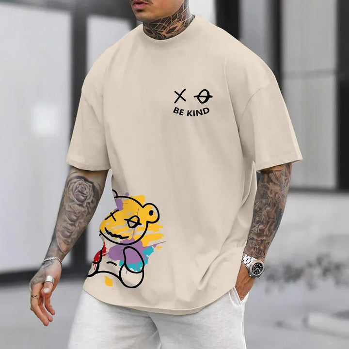 Beige oversized T-shirt with black text "BE KIND" and abstract colorful cartoon print on front worn by tattooed man