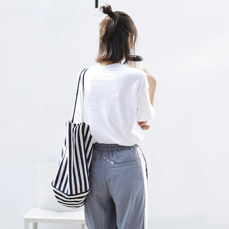 Woman in white t-shirt and gray pants with striped tote bag holding coffee cup against white background
