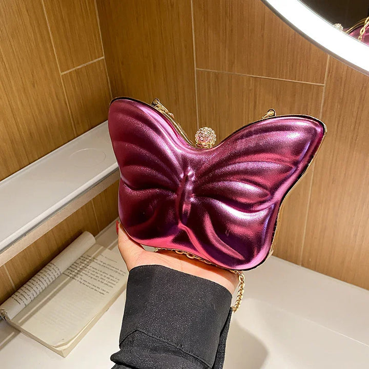 Hand holding shiny metallic purple butterfly-shaped clutch bag with gold chain in wooden room