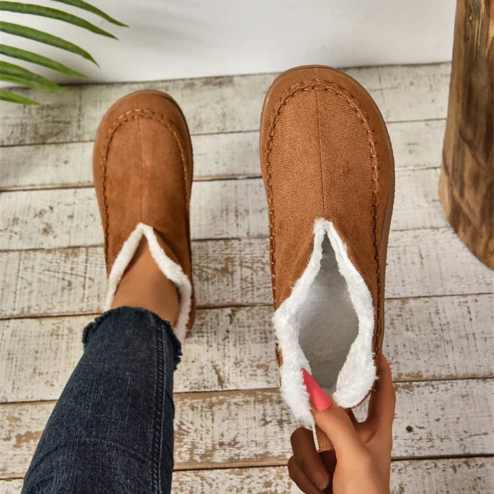 Brown suede slippers with white fleece lining on wooden floor, one worn and one held with pink nails