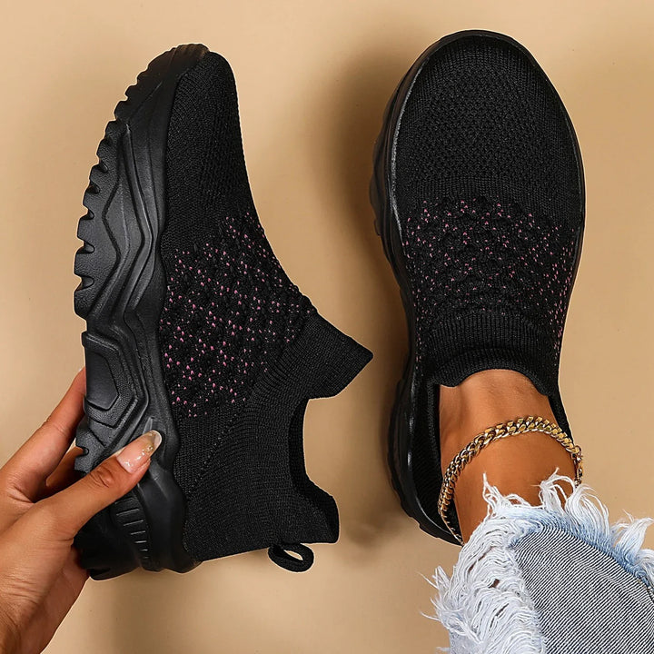 Black slip-on knit sneakers with chunky soles, pink speckles on upper, worn with frayed denim jeans and gold anklet