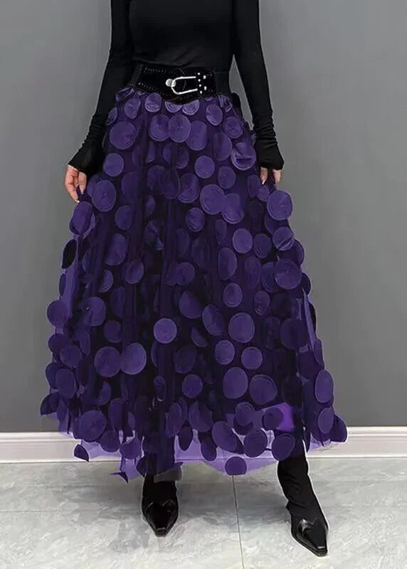Woman wearing a long purple skirt with large circular appliques, black belt, black top, and black pointed boots