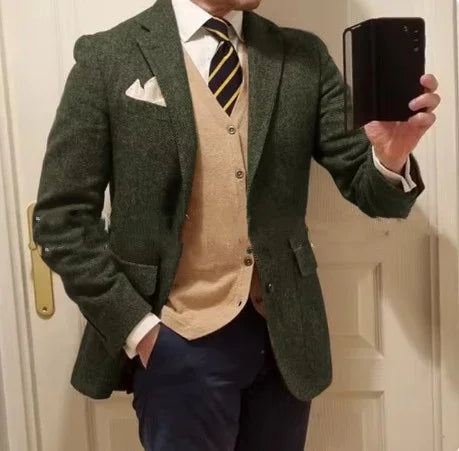 Man wearing green tweed blazer, beige cardigan, white shirt, striped tie, and navy pants taking mirror selfie