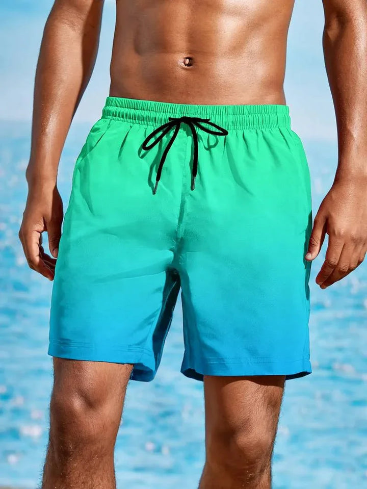 Men's green to blue gradient swim shorts with black drawstring, worn at the beach