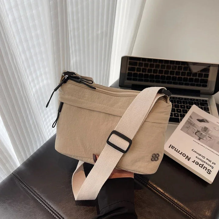 Beige crossbody bag with white adjustable strap held over black table next to laptop and book