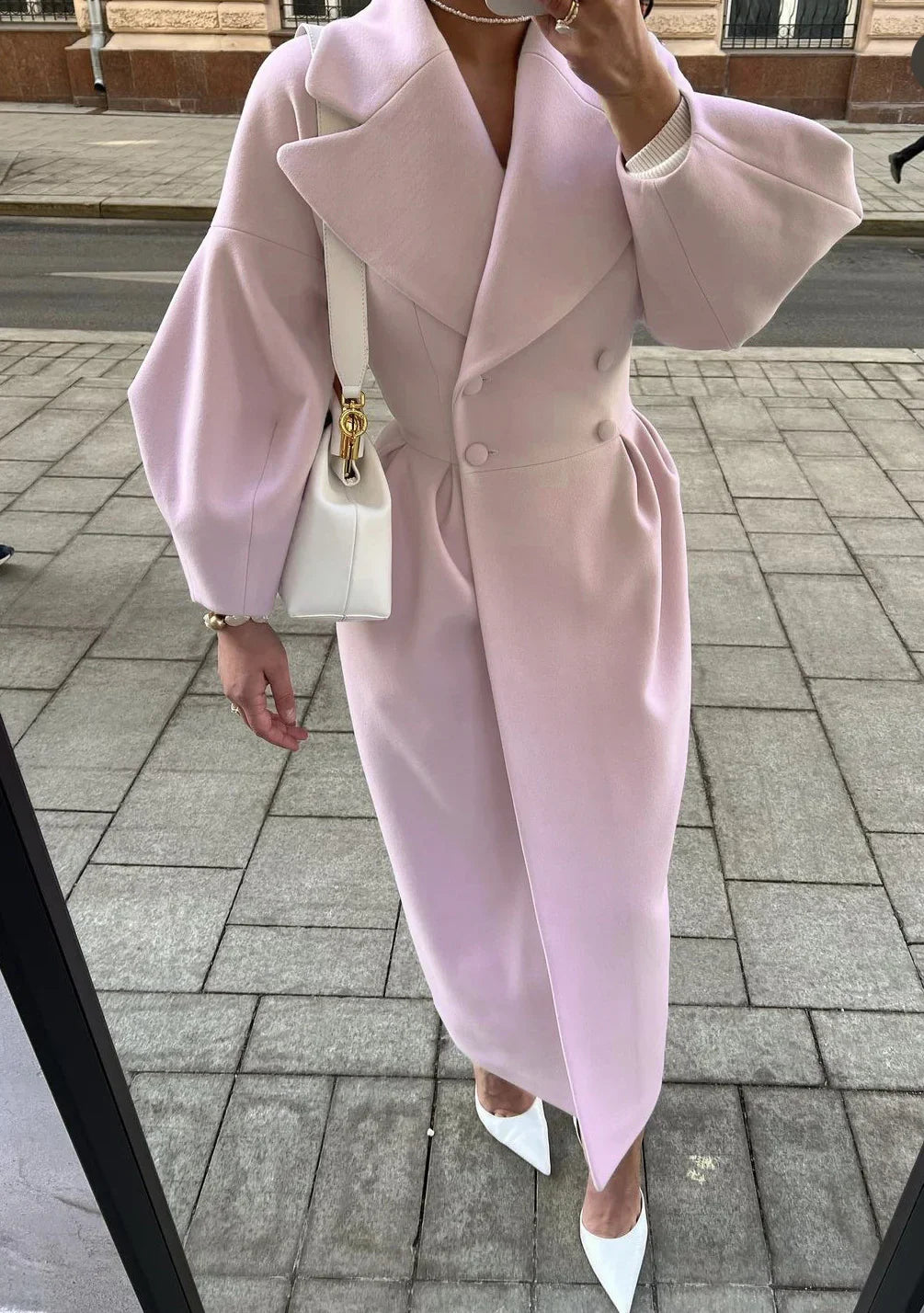 Woman wearing a light pink double-breasted coat with balloon sleeves, white pointed heels, and white handbag on city sidewalk