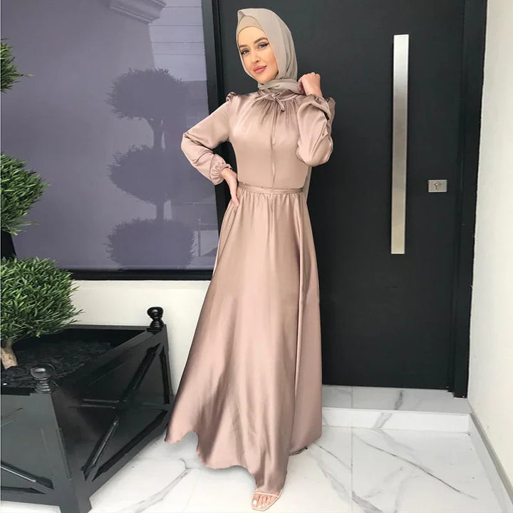 Woman in beige satin modest evening dress with matching hijab standing by black door and plants