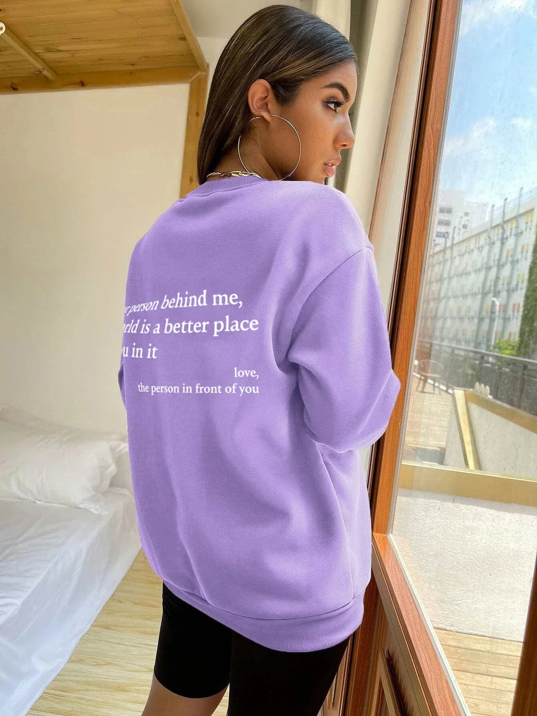 Young woman wearing oversized lavender sweatshirt with inspirational message, standing by window in bright room