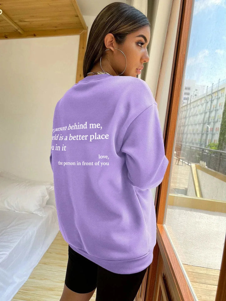 Young woman wearing oversized lavender sweatshirt with inspirational message, standing by window in bright room