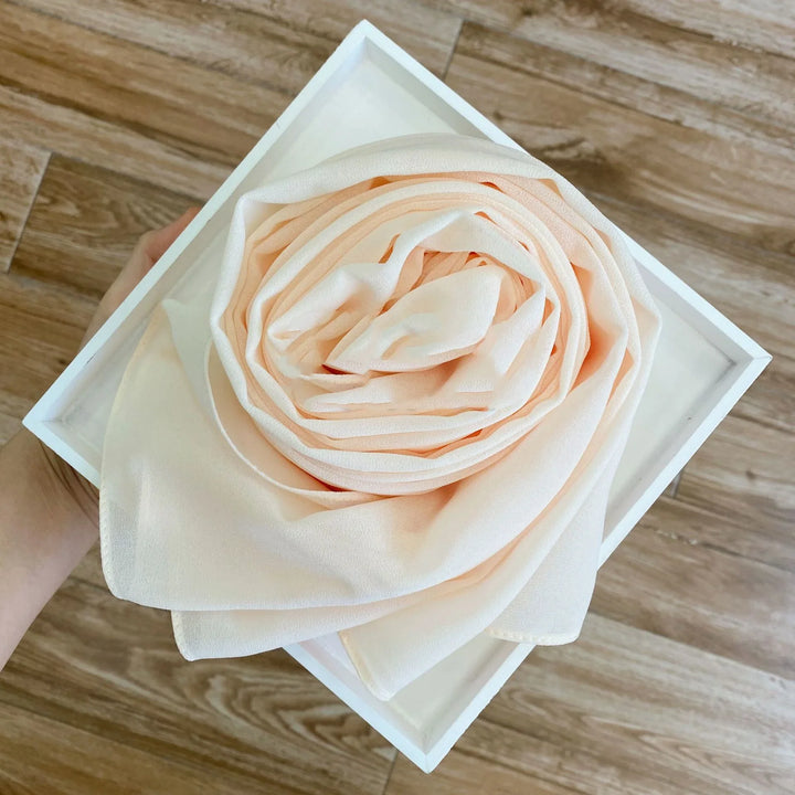 Hand holding white box with neatly folded light peach chiffon scarf resembling a rose on wooden floor