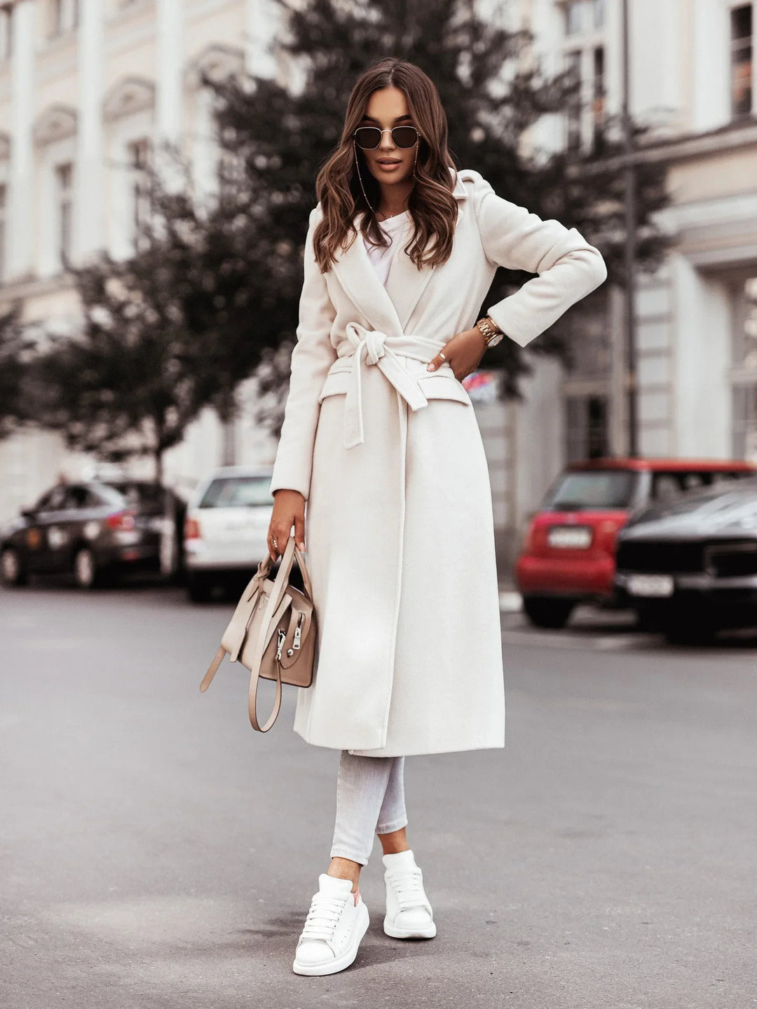 Fashionable woman in white belted wool coat, sunglasses, holding beige handbag on city street