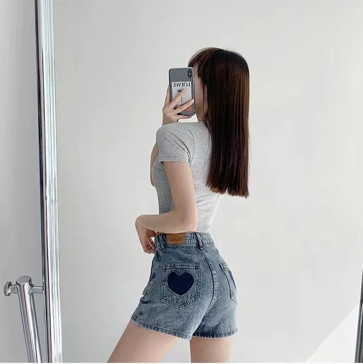 Woman taking a mirror selfie wearing gray fitted top and denim shorts with heart-shaped pocket detail