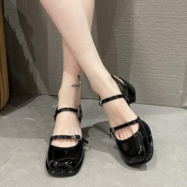 Close-up of legs wearing shiny black Mary Jane style block heel shoes with double straps on gray floor